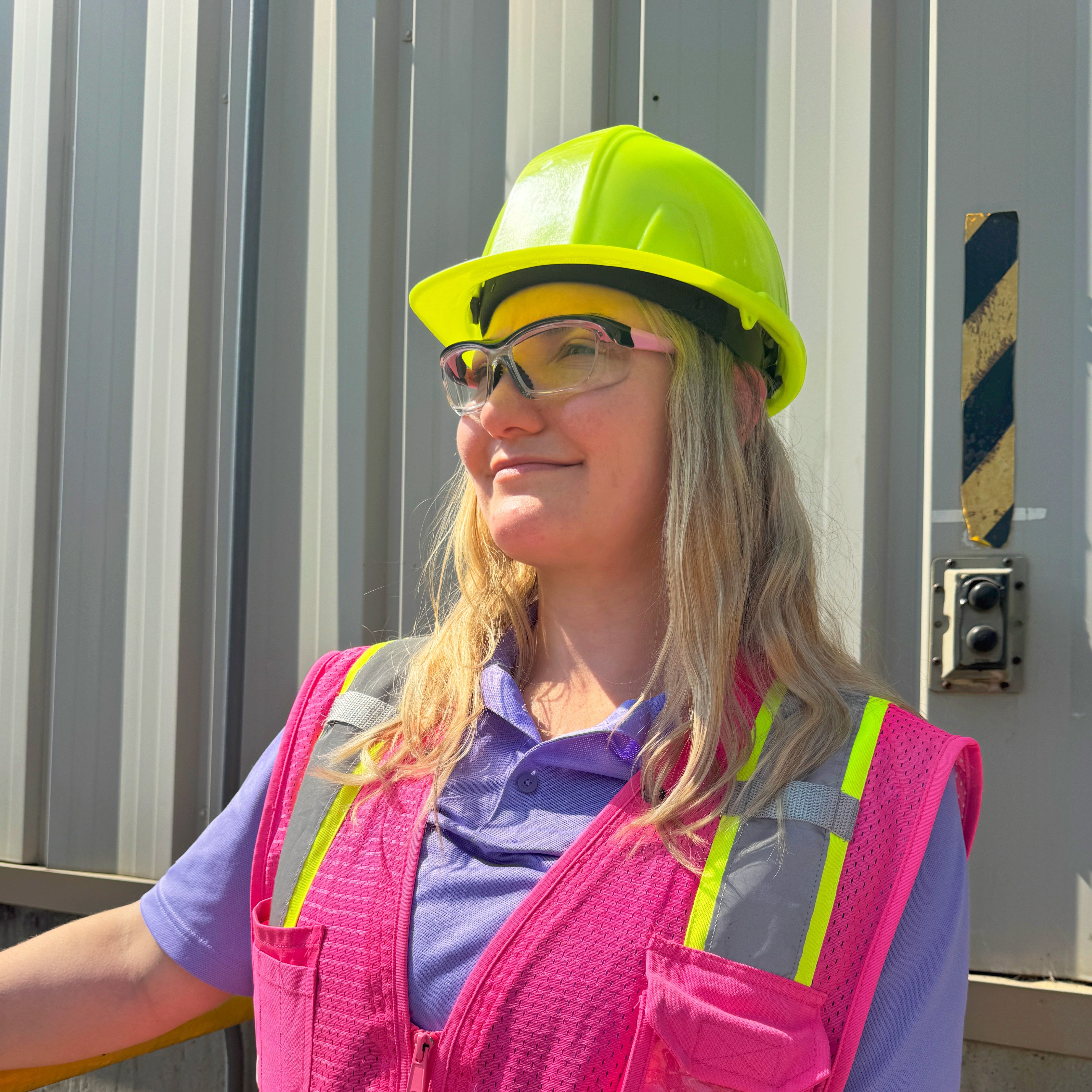 Prescription safety glasses in pink variant worn by woman in safety vest and hard hat outdoors